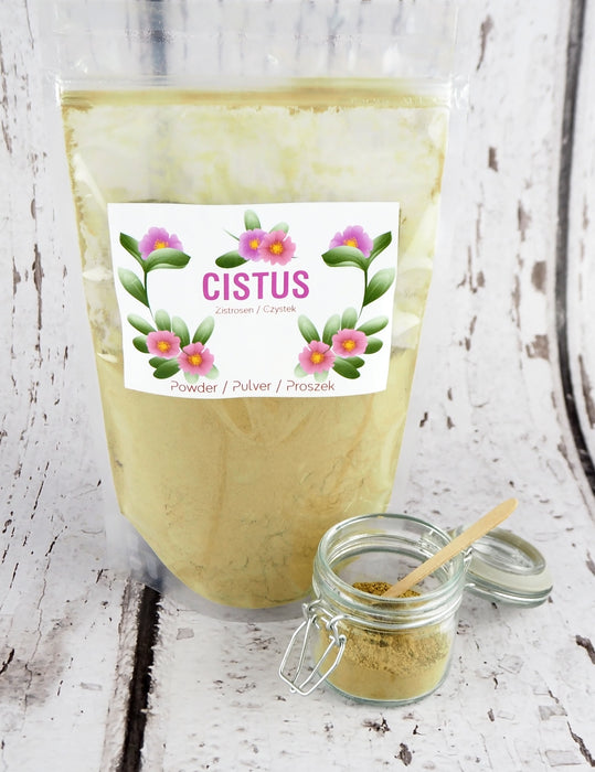 Cistus-Kräutertee in Pulverform in einer transparenten Tüte, präsentiert auf einem Holztisch neben einem offenen Glasgefäß mit Pulver und Holzlöffel.