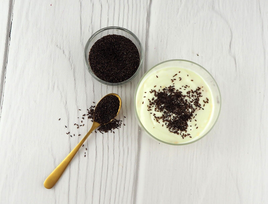 Schwarze Flohsamen auf einem goldenen Löffel neben einem Glas Joghurt mit Flohsamen bestreut auf weißem Hintergrund.