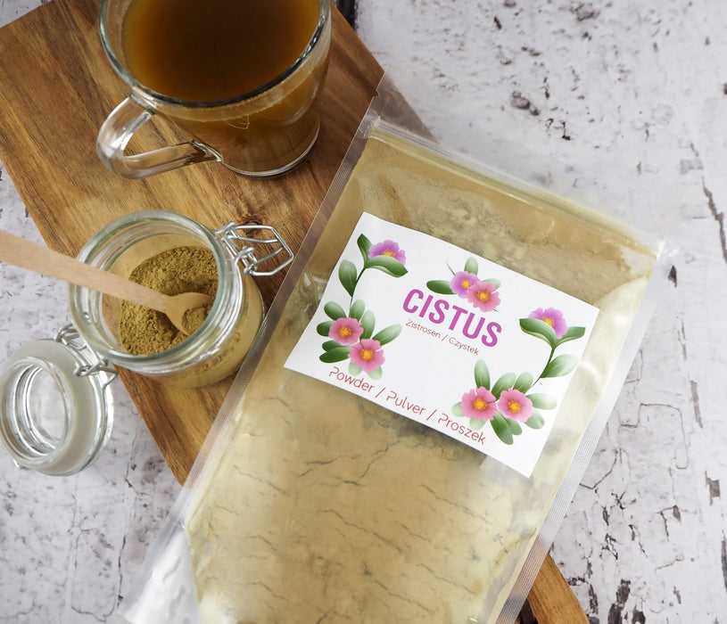 Cistus-Tee in Pulverform in einer durchsichtigen Verpackung, dargestellt auf einem Holzbrett mit einem Glas Tee und Holzlöffel, bereit zum Servieren.