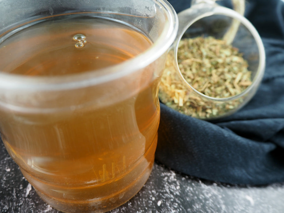 Das Bild zeigt eine Tasse mit klarem, goldfarbenem Tee, daneben ein Glas mit getrockneten, grünen Kräutern von Zitronenmyrte auf einem dunklen Untergrund mit einem schwarzen Tuch im Hintergrund.